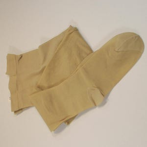 May include: A pair of beige tights, folded on a white surface. The tights are a solid, neutral colour, with a reinforced toe and a rolled waistband. The fabric appears to be a sheer nylon material, suitable for wear.
