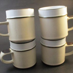 May include: Four cream-coloured ceramic mugs. Each mug has a white rim and a brown accent on the handle. The mugs are stacked in pairs, with two mugs on top of two others.