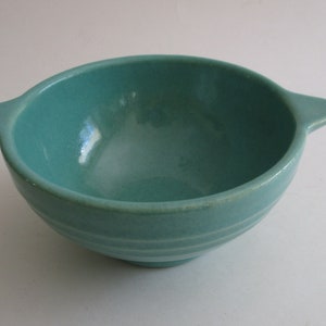 May include: A teal ceramic bowl with two small handles. The bowl has a smooth, glossy finish and a slightly textured surface. The bowl has a rounded shape with horizontal ridges near the base. The colour is a vibrant teal.