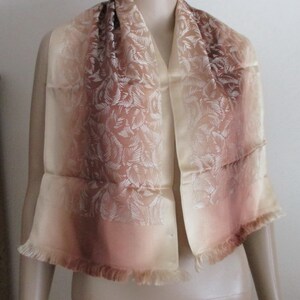 May include: A light brown silk scarf with a white floral pattern. The scarf has a gradient effect, fading from a darker brown to a lighter beige. The edges of the scarf are fringed.