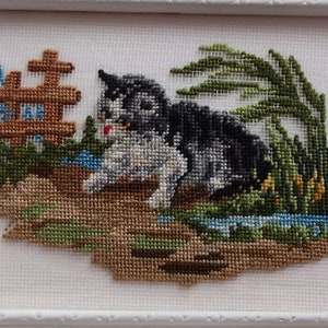 May include: Cross-stitch embroidery of a black and white cat sitting in a grassy area with a small wooden fence and a stream of water.
