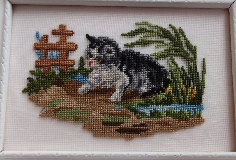 May include: Cross-stitch embroidery of a black and white cat sitting in a grassy area with a small wooden fence and a stream of water.