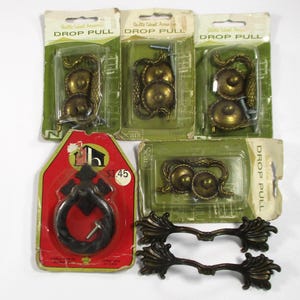 May include: A selection of vintage cabinet hardware, including drop pulls and decorative handles. The drop pulls are in their original packaging, with brass and black finishes. The packaging includes the text "DROP PULL". The price tag reads "£1.45".