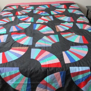 Peut inclure: Une couverture noire avec un motif de ventilateur répété en rouge, bleu, rose et vert. La couverture est posée sur un lit, mettant en valeur le motif coloré. Les formes de ventilateur sont disposées en grille.