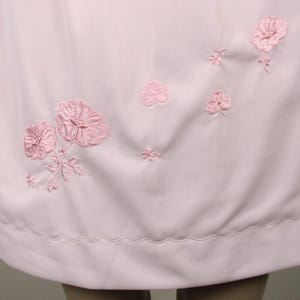 May include: A pale pink dress with embroidered pink floral details. The dress has a scalloped hemline and various sizes of embroidered flowers across the fabric.