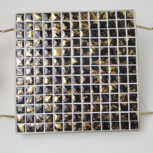 May include: Square trivet with a mosaic tile design. The tiles are black and gold, arranged in a grid pattern. It has a gold-coloured metal frame and handles.
