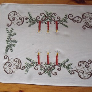 Peut inclure: Un chemin de table blanc avec un motif brodé de couronne de Noël avec des bougies rouges et des accents dorés.