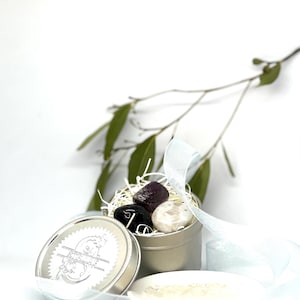 May include: A small, silver tin with a lid, containing three polished stones in shades of purple, black, and white, nestled in straw. A small white dish with crystals and a sprig of greenery are also present.