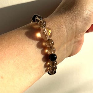 May include: A beaded bracelet featuring alternating round, translucent smoky quartz beads and opaque black beads. One clear bead contains a small, metallic gold-coloured decorative element inside. The beads are strung together in a single circular row.