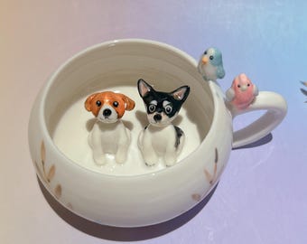May include: White ceramic mug with a round bowl shape, featuring two small dog figurines inside. The mug's handle has two bird figurines. The mug has gold leaf accents. The dogs are a Jack Russell Terrier and a Chihuahua.