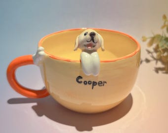 May include: A ceramic coffee mug with a light yellow interior and exterior, featuring an orange handle. A small, smiling dog figurine is perched on the rim. The name "Cooper" is written on the side of the mug.