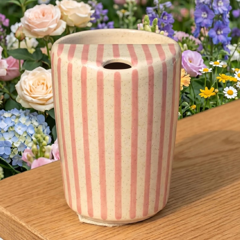 Pu&ograve; includere: Un vaso cilindrico in ceramica con base color crema e strisce rosa verticali. Il vaso ha un unico foro nella parte superiore ed &egrave; posto su una superficie di legno. Lo sfondo presenta una variet&agrave; di fiori colorati.