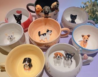 May include: A collection of ceramic mugs, each featuring a different dog or cat figurine inside. The mugs are in various colors, including white, pink, and yellow. Some mugs have paw prints or heart designs.