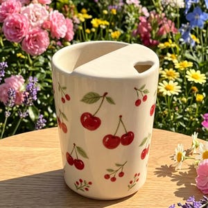 May include: A cream-colored ceramic tumbler featuring a repeating pattern of red cherries with green leaves and stems. The cup has a flat lid with a small triangular opening for drinking, and the exterior surface is smooth with a speckled finish.