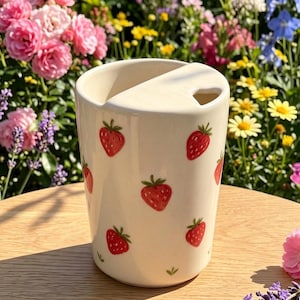 May include: A cream-colored ceramic tumbler featuring a repeating pattern of small red strawberries with green leaves. The top of the tumbler has a partial cover with a small heart-shaped cutout, designed for holding a straw or utensil.