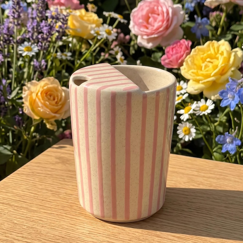 Pu&ograve; includere: Una tazza in ceramica beige con strisce rosa verticali e un coperchio abbinato. La tazza cilindrica &egrave; appoggiata su una superficie di legno. Lo sfondo presenta una variet&agrave; di fiori colorati, tra cui rose e margherite.