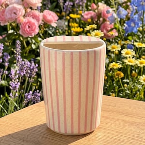 Pu&ograve; includere: Un vaso in ceramica color crema con strisce rosa verticali. Il vaso ha una forma rettangolare con una parte superiore curva. Il vaso &egrave; appoggiato su una superficie di legno, con un colorato giardino fiorito sullo sfondo.