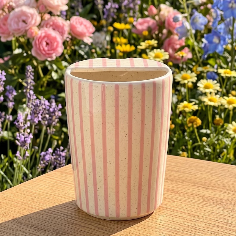 Pu&ograve; includere: Un vaso in ceramica color crema con strisce rosa verticali. Il vaso ha una forma rettangolare con una parte superiore curva. Il vaso &egrave; appoggiato su una superficie di legno, con un colorato giardino fiorito sullo sfondo.