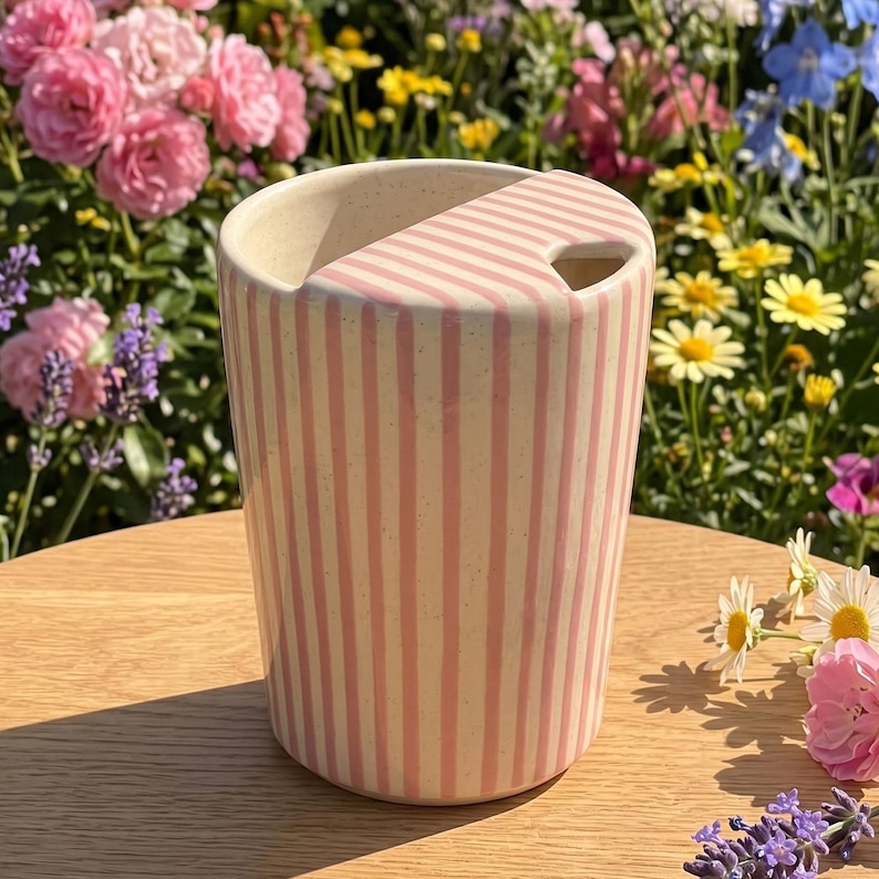 Pu&ograve; includere: Un bicchiere in ceramica con un motivo a righe verticali rosa e crema. Il bicchiere ha un manico curvo e una base arrotondata. Lo sfondo presenta un tavolo di legno e un giardino con fiori colorati.