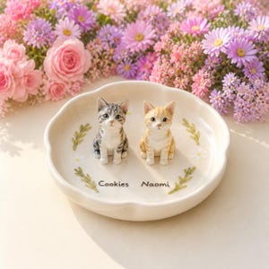 May include: A cream-coloured ceramic dish with scalloped edges featuring two small cat figurines, one grey tabby and one orange tabby. The dish is decorated with painted green sprigs and white daisies, with the names Cookies and Naomi printed.