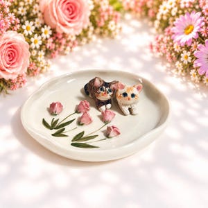 May include: A round, off-white ceramic trinket dish featuring two small, sculpted cat figurines—one brown tabby and one cream-colored—resting beside hand-painted pink tulip buds with green stems and leaves on the interior surface.