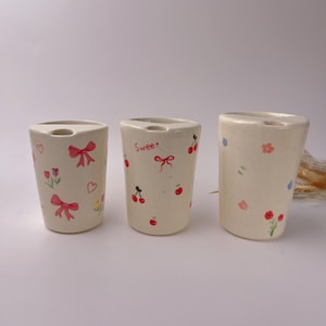 May include: Three off-white ceramic toothbrush holders with hand-painted designs. One features pink bows, hearts, and flowers. Another has cherries and the word "Sweet". The third has flowers. Each has a top slot.