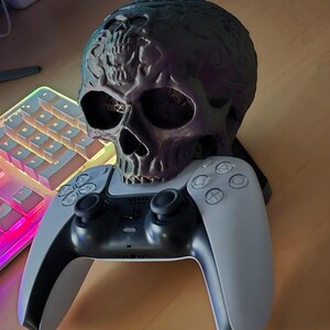 May include: A black and purple skull-shaped stand holds a white PlayStation 5 controller. The controller is on a wooden surface next to a white keyboard with rainbow-colored lights.
