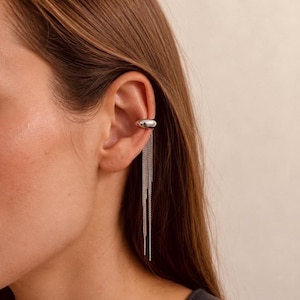Könnte beinhalten: Ein silberfarbener Ear Cuff mit einem glatten, polierten und abgerundeten Band, das auf dem Knorpel sitzt. An der Unterseite des Cuffs sind mehrere feine, dünne Silberketten befestigt, die vertikal vom Ohr herabhängen.