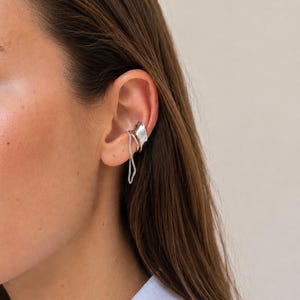 Könnte beinhalten: Ein silberfarbener Ohrring bestehend aus einer glatten, geschwungenen Metallplatte, die den oberen Knorpel umschließt, verbunden durch eine zarte, dünne Silberkette, die nach unten zum Ohrläppchen führt und einen mehrlagigen Effekt erzeugt.