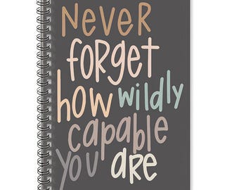 May include: A spiral-bound notebook with a dark gray cover. The cover features the words "Never Forget How Wildly Capable You Are" in various pastel colors. The notebook has a silver spiral binding.