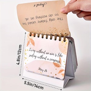 May include: A small desktop calendar with a white base and a pink and brown cover. The calendar features a quote: "Living without an aim is like sailing without a compass." The date is May 26. The calendar measures 4.8 inches tall and 5.5 inches wide.