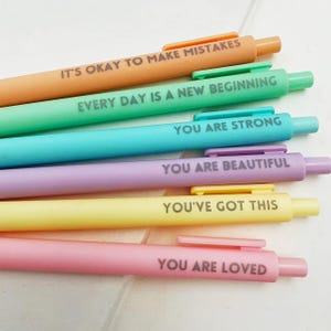 May include: Six colourful pens in orange, green, blue, purple, yellow, and pink. Each pen has an inspirational message printed on it, such as "EVERY DAY IS A NEW BEGINNING" and "YOU ARE BEAUTIFUL".