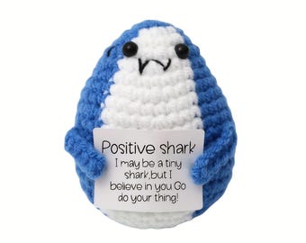 May include: A handmade blue and white crocheted shark plushie holding a white sign. The sign reads "Positive shark, I may be a tiny shark, but I believe in you Go do your thing!" The shark has black eyes and a small mouth.