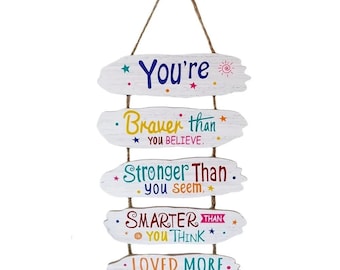 May include: A wooden wall hanging with five white, oval-shaped planks. Each plank has a motivational phrase in colorful text: "You're", "Braver than you believe", "Stronger than you seem", "Smarter than you think", and "Loved more than you realize". The planks are connected by twine.