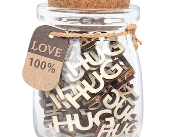 May include: A clear glass jar with a cork stopper filled with small wooden cutouts of the word HUG. A brown paper tag is tied to the neck of the jar with twine, featuring the text LOVE and 100% printed in dark brown ink on the front surface.