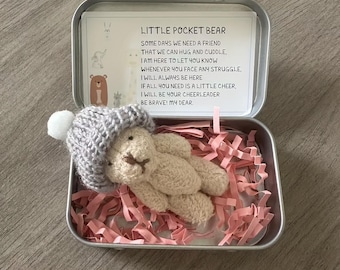 May include: A small, beige crocheted bear wearing a gray hat with a white pom-pom, nestled in a metal tin filled with pink paper shreds. The tin contains a card with the text "LITTLE POCKET BEAR" and a comforting message.