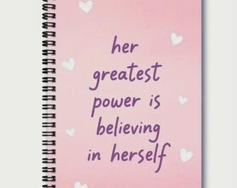 May include: Pink spiral-bound notebook with the text "her greatest power is believing in herself" in purple script. White heart shapes are scattered across the cover. The notebook's binding is black.