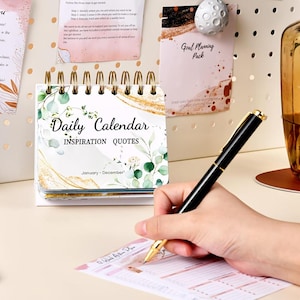 May include: A small, white desktop calendar with a gold spiral binding. The front cover features a watercolor design of green leaves and gold accents. The calendar is titled "Daily Calendar Inspiration Quotes" and includes the months January through December.