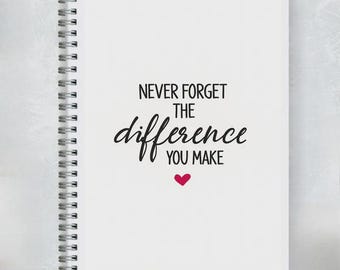 May include: White spiral-bound notebook with the text "NEVER FORGET THE difference YOU MAKE" in black script and a small red heart. The notebook is on a light gray background.