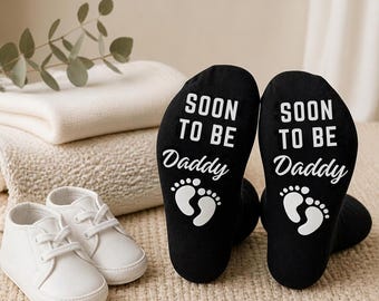 May include: A pair of black socks with white text on the soles that reads SOON TO BE Daddy, accompanied by a graphic of two small baby footprints. Next to the socks is a pair of small white baby shoes with white laces and a rounded toe design.