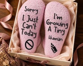 May include: A pair of light pink fuzzy socks inside a gift box. The left sock features the black text Sorry I Just Can't Today and a prohibition symbol. The right sock features the black text I'm Growing A Tiny Human and a baby bottle icon.