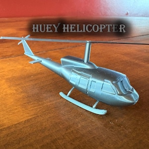 May include: A silver-colored model helicopter with the text "HUEY HELICOPTER" above it. The helicopter has a main rotor, tail rotor, and landing skids. The model is sitting on a wooden surface.