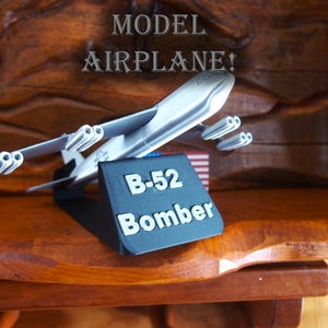 May include: A silver model airplane on a black stand with the text "B-52 Bomber". The airplane is on a wooden surface with the text "MODEL AIRPLANE!" above it.