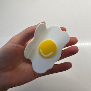 May include: A white plastic fried egg shaped phone holder with a yellow yolk and a white smiley face.