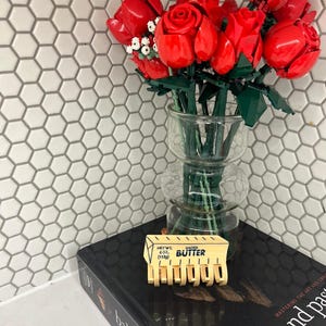 May include: A bouquet of red roses constructed from plastic building bricks arranged in a clear glass vase. A yellow plastic hair claw clip shaped like a stick of salted butter with the text NET WT. 4 OZ. (113g) BUTTER is placed in front.