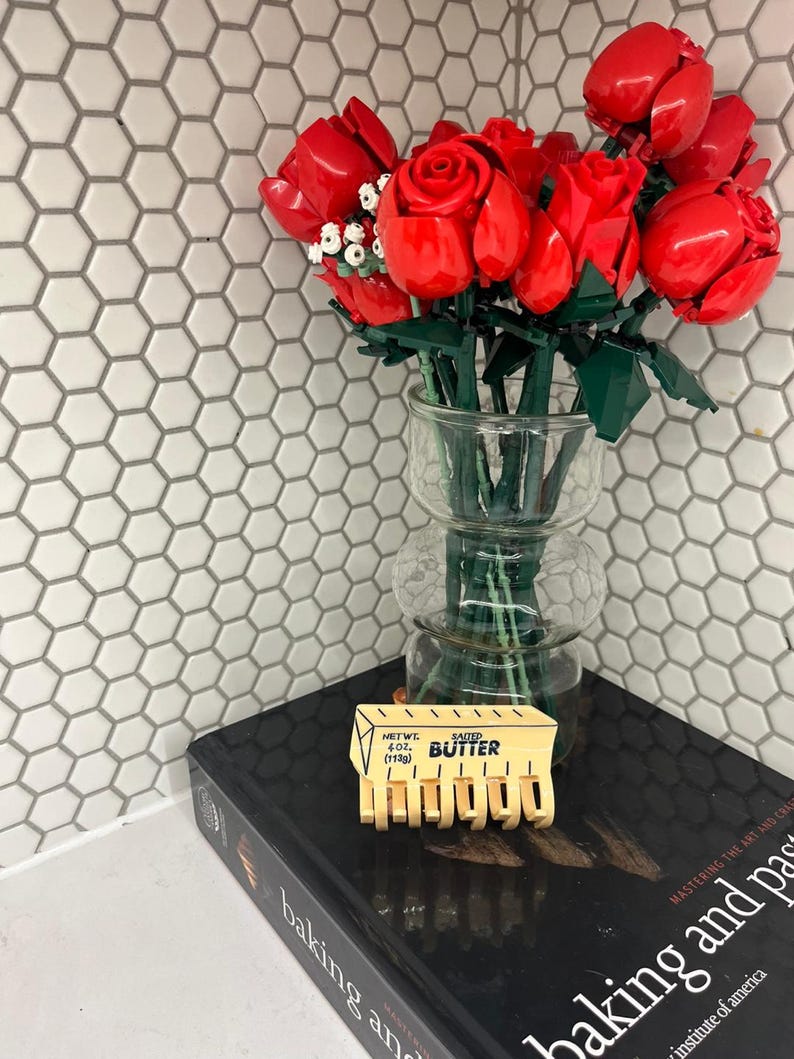 May include: A bouquet of red roses constructed from plastic building bricks arranged in a clear glass vase. A yellow plastic hair claw clip shaped like a stick of salted butter with the text NET WT. 4 OZ. (113g) BUTTER is placed in front.