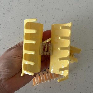 May include: A pale yellow plastic hair claw clip with a glossy finish, featuring a central metal spring mechanism and interlocking teeth on both sides to secure hair in place. The design is simple, functional, and lightweight for daily use.