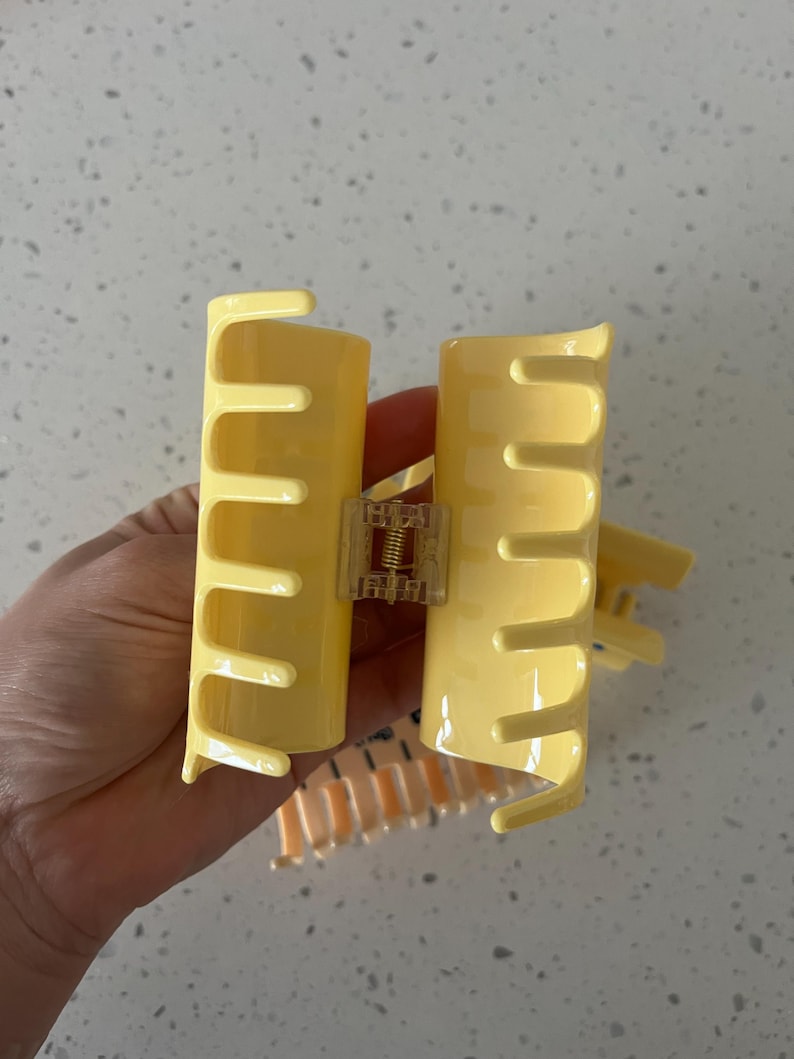 May include: A pale yellow plastic hair claw clip with a glossy finish, featuring a central metal spring mechanism and interlocking teeth on both sides to secure hair in place. The design is simple, functional, and lightweight for daily use.