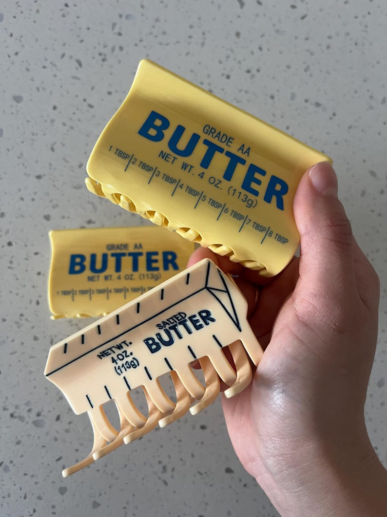 May include: Two yellow hair claw clips shaped like butter sticks. The top clip features blue text reading BUTTER, GRADE AA, NET WT. 4 OZ. (113g) and tablespoon markings. The bottom clip features black text reading SALTED BUTTER, NET WT. 4 OZ. (113g).
