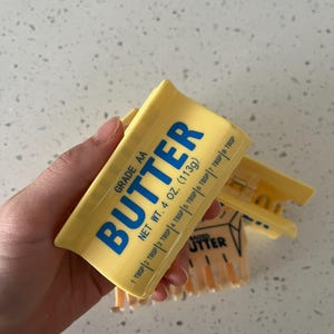 May include: A yellow plastic hair claw clip designed to resemble a stick of butter. The clip features blue text that reads GRADE AA BUTTER, NET WT. 4 OZ. (113g), and includes measurement markings from 1 TBSP to 8 TBSP along the side of the clip.
